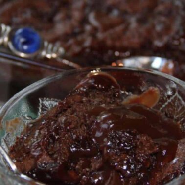 A glass bowl filled with timeless chocolate dessert sits in front, a spoon nestled within. Behind, a larger dish brims with more of this classic treat, resting on a checkered tablecloth.