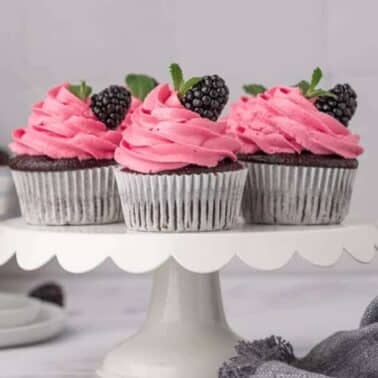 Chocolate cupcakes with pink frosting and blackberries on a white cake stand, with a milk bottle and bowl of blackberries in the background.