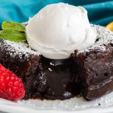 A chocolate lava cake with molten center, topped with whipped cream and mint, is on a white plate. Raspberries and a gold spoon are beside the cake.