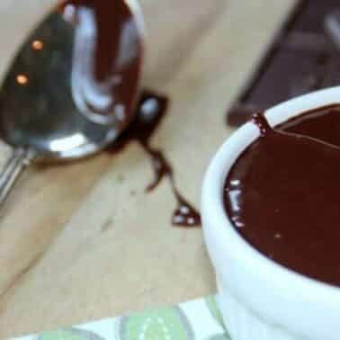 A bowl of chocolate nutella sauce next to a spoon with chocolate nutella sauce on it, resting on a wooden surface with a chocolate bar nearby.