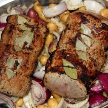 Roasted pork roasts with bay leaves, surrounded by potatoes, red onions, and garlic in a baking pan.
