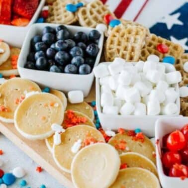 A patriotic-themed charcuterie breakfast board with strawberries, blueberries, marshmallows, waffles, pancakes, cherries, and colorful candies on a USA flag backdrop.
