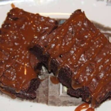 Two chocolate brownies drizzled with caramel and sprinkled with sea salt on a decorative plate.