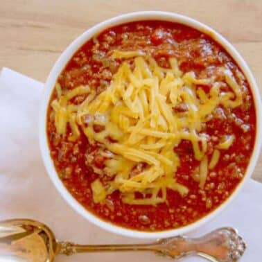 A bowl of chili topped with shredded cheese on a white napkin, accompanied by a silver spoon.