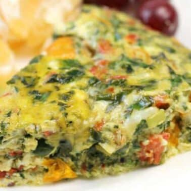 A slice of crustless frittata with visible spinach, tomatoes, and cheese, served on a plate with slices of kiwi, orange, and a small bunch of red grapes.