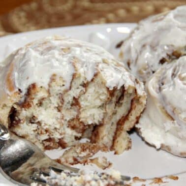 Three cinnamon rolls with icing on a white plate, one partially eaten with a fork nearby.
