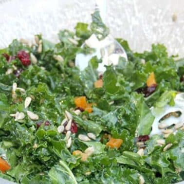 A glass bowl of kale salad with dried fruits, sunflower seeds, and two serving utensils.