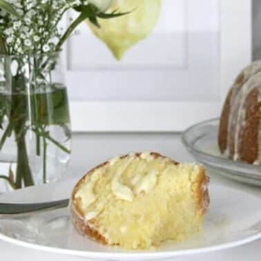 Slice of lemon pound cake with icing on a white plate, next to a fork, with a whole cake in the background. A vase with flowers and a framed lemon picture are on the table.