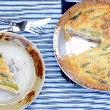 A quiche with a slice removed on a striped cloth next to a plate with the slice and a fork and knife.