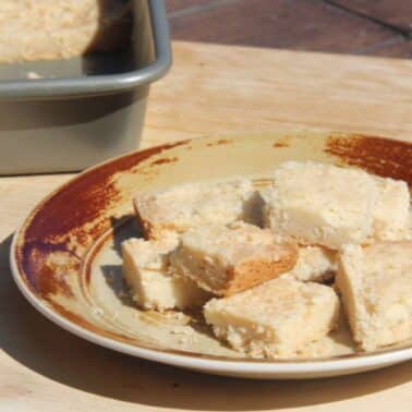 A brown-rimmed plate on a wooden surface holds several pieces of crumbly shortbread cookies. A baking tray with more shortbread cookies is in the background.