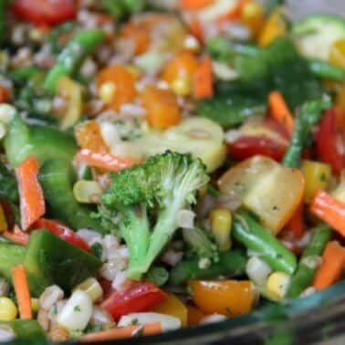A glass bowl filled with a colorful mixed vegetable salad, including farro, broccoli, bell peppers, carrots, corn, and zucchini.