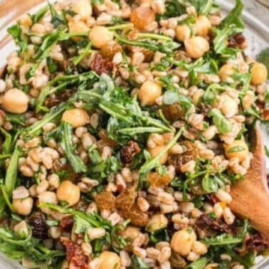 A glass bowl filled with a salad of arugula, chickpeas, farro, raisins, and sun-dried tomatoes, being tossed with wooden salad forks.