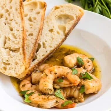 A bowl with French Quarter shrimp in a seasoned sauce, garnished with herbs, and three slices of rustic bread on the side.