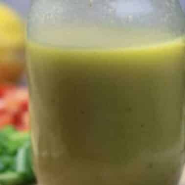 A glass jar filled with French salad dressing, is in focus. In the background, blurred vegetables and a lemon are visible.