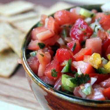 A brown bowl filled with fresh salsa containing diced tomatoes, onions, peppers, and cilantro. In the background, there are square crackers on a wooden surface.