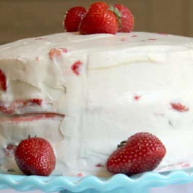 A round frosted strawberry cake with strawberries on top and around the base, set on a light blue cake stand.