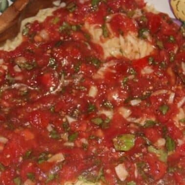 Plate of spaghetti topped with chunky red tomato sauce, onions, and herbs, beside a wooden spoon.