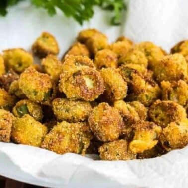 A plate of fried okra pieces on a paper towel-lined dish, with green herbs in the background.