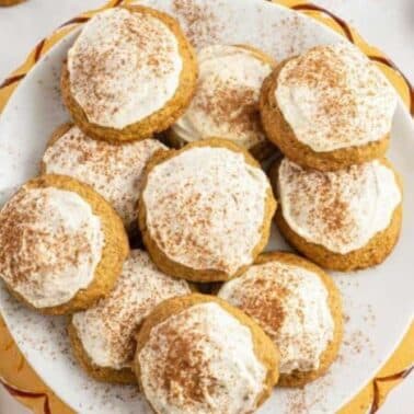 A plate of frosted pumpkin cookies sprinkled with cinnamon, placed on a white and gold-rimmed plate. A few cookies are scattered around the plate on a white surface.