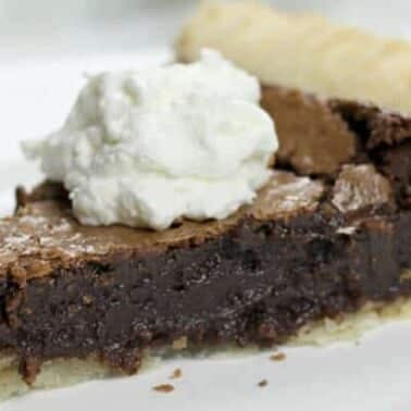A slice of fudge pie with a flaky crust, topped with a dollop of whipped cream, on a white plate.