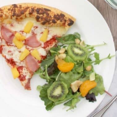 A slice of pizza with ham and pineapple next to a salad with kiwi, mandarin, and greens on a white plate. A fork rests on a gray napkin beside the plate.
