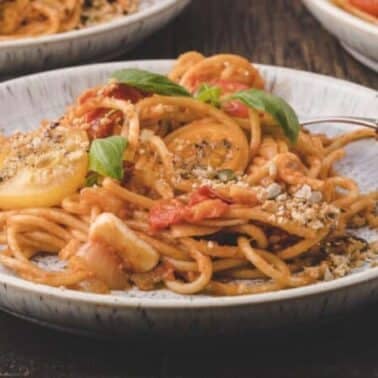 A plate of spaghetti with heirloom tomato sauce, garnished with sliced vegetables, herbs, and sprinkled nuts, on a wooden table.