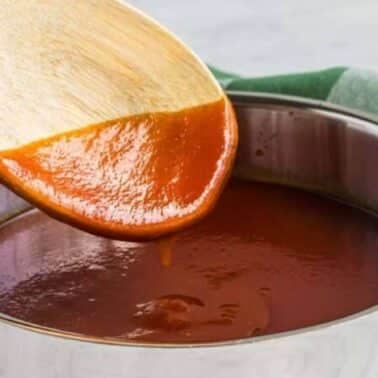 Wooden spoon with BBQ sauce over a stainless steel pot, placed on a woven mat, with a green and white cloth nearby.