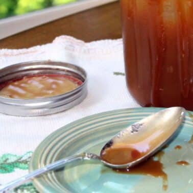 Jar of caramel sauce with a lid beside it on a cloth, a spoon and drips on a small plate nearby.