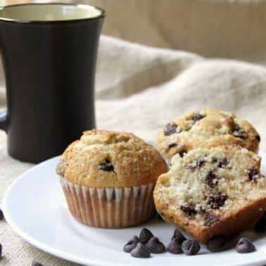 Chocolate chip muffins on a plate with scattered chips, next to a black mug, set on a beige fabric background.