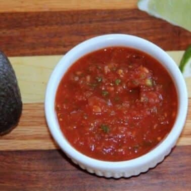 A bowl of red homemade salsa next to a whole avocado and two lime wedges on a wooden surface.