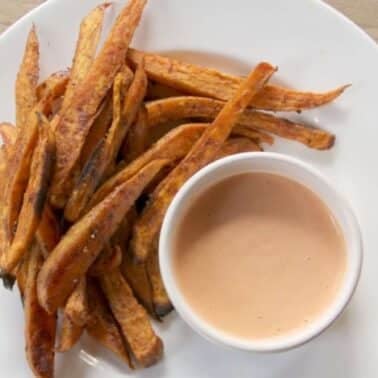A plate of sweet potato fries next to a small bowl of dipping sauce on a wooden surface.