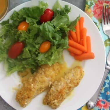 A plate with two breaded chicken tenders, a green salad with cherry tomatoes, and baby carrots. A fork, knife, and colorful napkin are placed beside the plate. A drink is in the background.