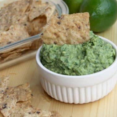 Bowl of guacamole with a tortilla chip, surrounded by more chips and a lime on a wooden surface.