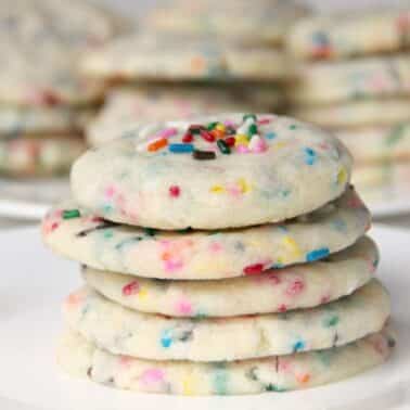 A stack of funfetti sugar cookies with colorful sprinkles on a white plate, with more cookies in the background.