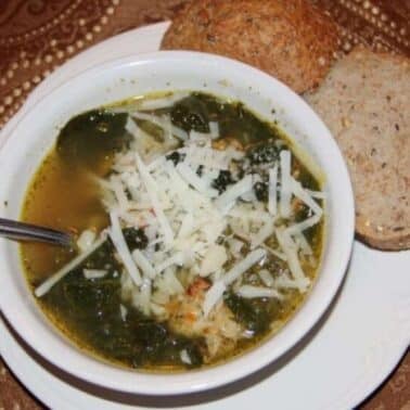 A bowl of Italian wedding soup with greens and shredded cheese, placed on a white saucer with a spoon. Two slices of whole-grain bread are on the side. The background is a patterned brown fabric.