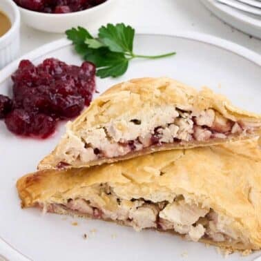 A plate with sliced pastry pie filled with turkey and cranberries, accompanied by cranberry sauce and parsley. A small dish of gravy and cranberries are in the background.
