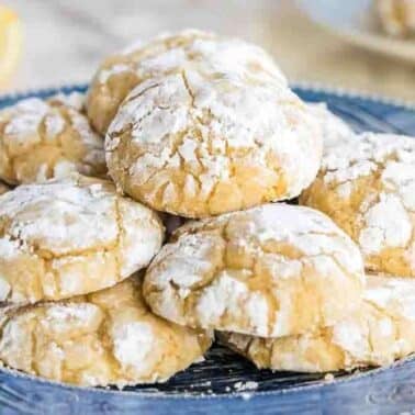 A plate of lemon crinkle cookies dusted with powdered sugar is arranged on a table.