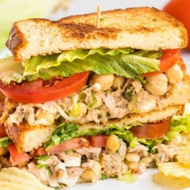 A stacked sandwich with tuna salad, lettuce, tomato, and filling on toasted bread, garnished with potato chips on a white plate.