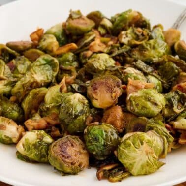 A white plate filled with roasted maple and balsamic glazed Brussels sprouts mixed with small pieces of bacon.