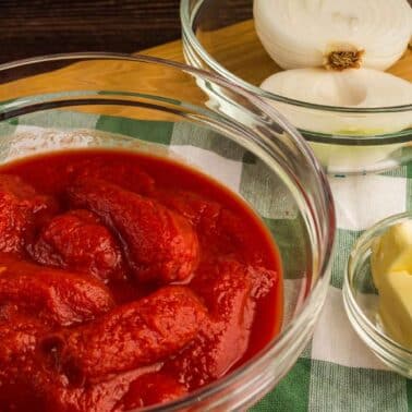 A glass bowl of whole peeled tomatoes in tomato sauce, next to glass bowls containing butter cubes and a halved onion, placed on a green and white checkered cloth on a wooden surface.