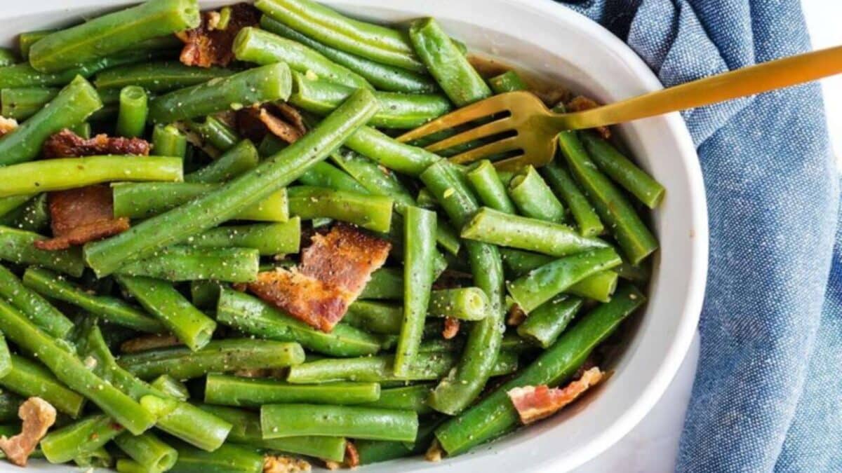 A white dish filled with seasoned green beans and pieces of bacon, placed next to a blue cloth. A gold fork rests in the dish.