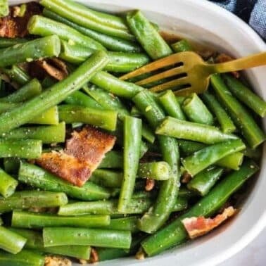 A white dish filled with seasoned green beans and pieces of bacon, placed next to a blue cloth. A gold fork rests in the dish.