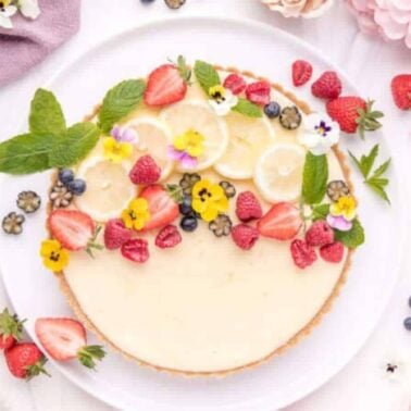 A timeless dessert, the cheesecake is adorned with lemon slices, strawberries, raspberries, blueberries, and edible flowers on a white plate. Surrounding are loose fruits, flowers, and a light purple cloth.