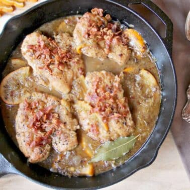 Chicken breasts cooked in a cast iron skillet with bacon bits and sauce, garnished with orange slices and a bay leaf. Spoon and napkin are beside the skillet.