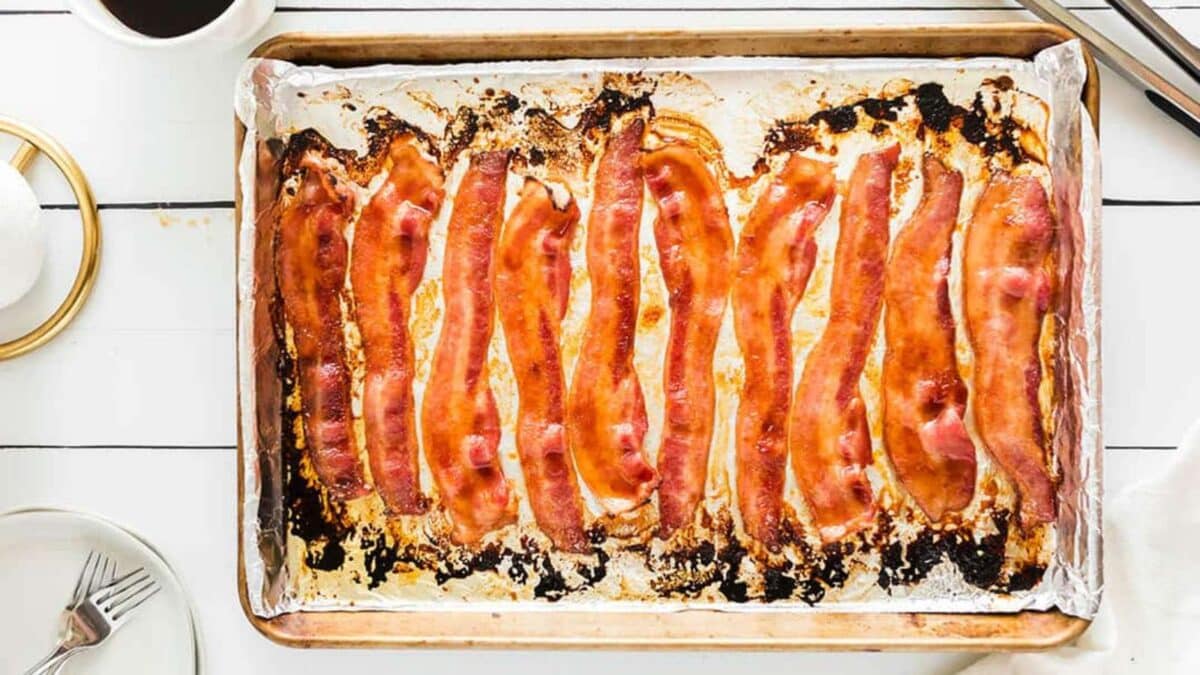 Ten strips of baked bacon on a foil-lined baking sheet, with a small fork on a plate and a cup nearby.
