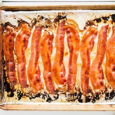 Ten strips of baked bacon on a foil-lined baking sheet, with a small fork on a plate and a cup nearby.