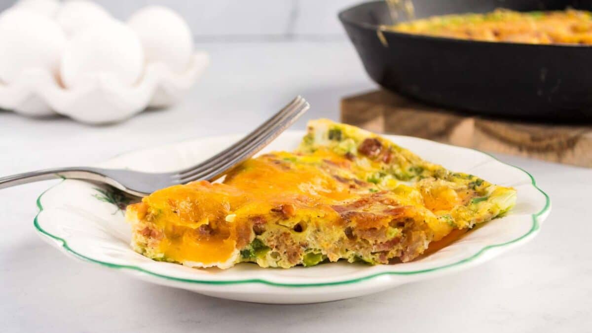 A slice of an oven baked omelet with cheese and greens on a white plate with a fork, with a skillet and eggs in the background.