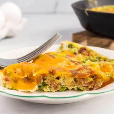 A slice of an oven baked omelet with cheese and greens on a white plate with a fork, with a skillet and eggs in the background.