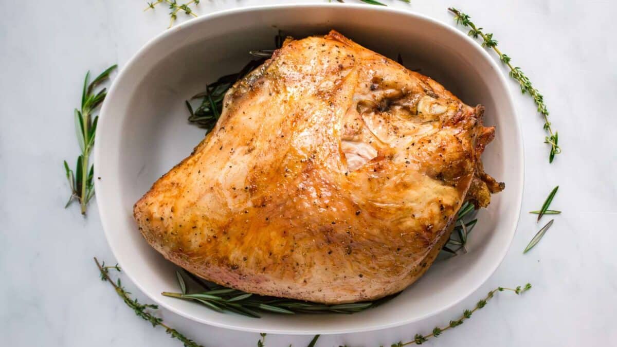 Roasted turkey breast in a white oval dish, garnished with fresh rosemary and thyme sprigs on a white marble surface.