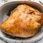 Roasted turkey breast in a white oval dish, garnished with fresh rosemary and thyme sprigs on a white marble surface.